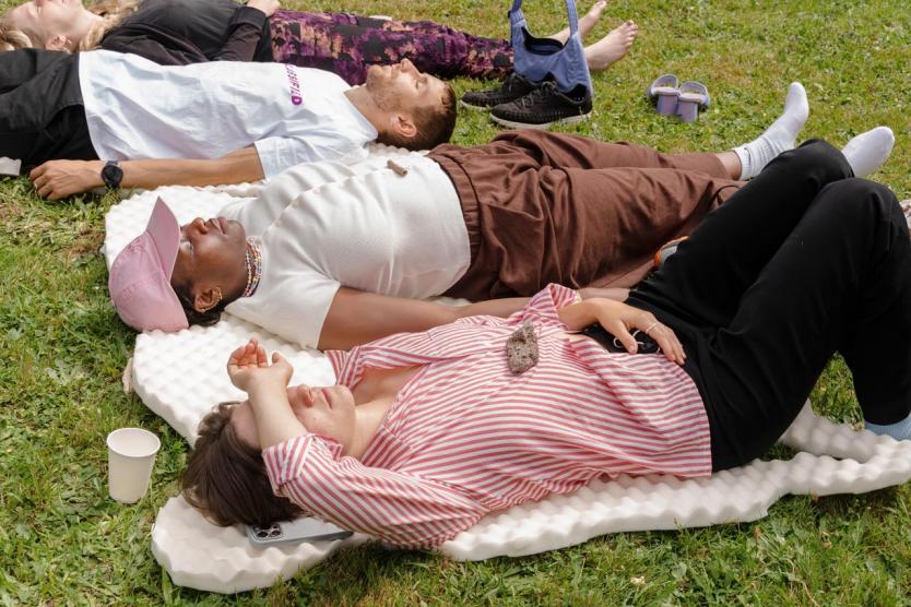 Mehrere Personen liegen entspannt auf Schaumstoffmatten im Gras, teils mit geschlossenen Augen, barfuß oder mit Socken, persönliche Gegenstände und Becher um sie herum verstreut.