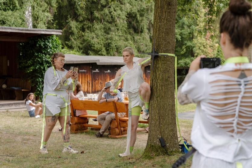 Eine Person steht mit erhobenem Bein an einen Baum gelehnt, festgebunden mit neonfarbenen Bändern, während zwei weitere Personen die Szene mit Kameras und Smartphones aufnehmen; im Hintergrund sitzen Menschen an Holzbänken im Grünen.