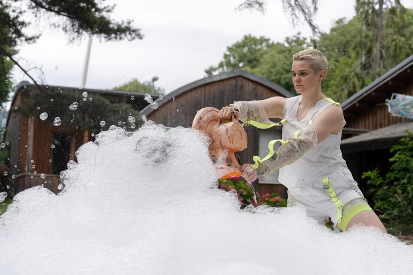 Schaum strömt in dichten Wellen aus einem orangefarbenen Gebläse, das über einem Becken gehalten wird – umgeben von grüner Kulisse und Holzbauten.