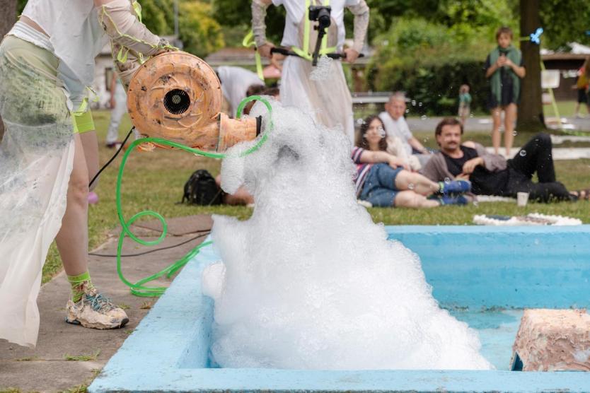 Aus einem skulptural gestalteten Gebläse strömt Schaum in ein türkisblaues Becken, während im Hintergrund Menschen entspannt auf der Wiese sitzen und das Geschehen beobachten.