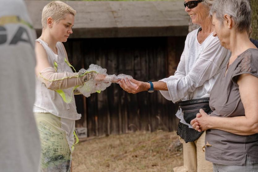 Eine Person mit kurzem blondem Haar reicht einer älteren Frau eine Hand voller Schaum, beide lächeln, daneben steht eine weitere beobachtende Frau – Begegnung im Freien vor dunkler Holzwand.