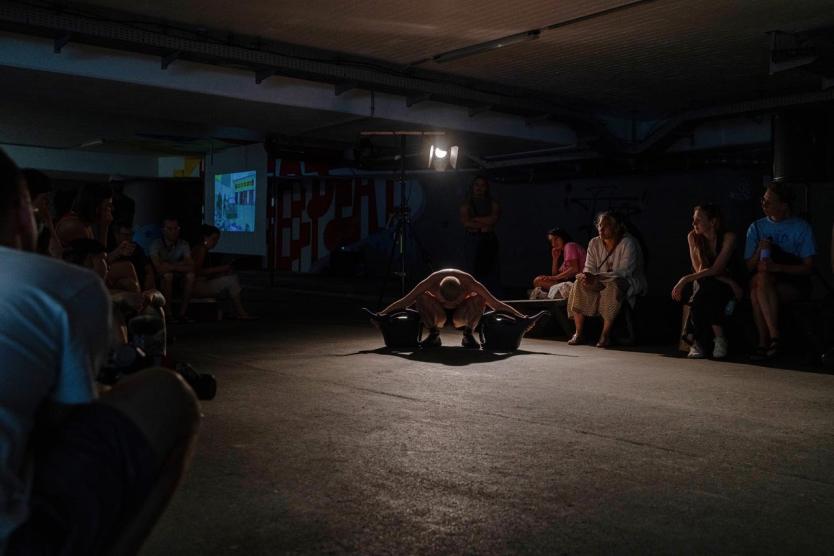 Eine Person hockt im Scheinwerferlicht auf dem Boden eines Parkhauses, mit je einer Gießkanne in der Hand, während ringsum ein sitzendes Publikum die Szene beobachtet.