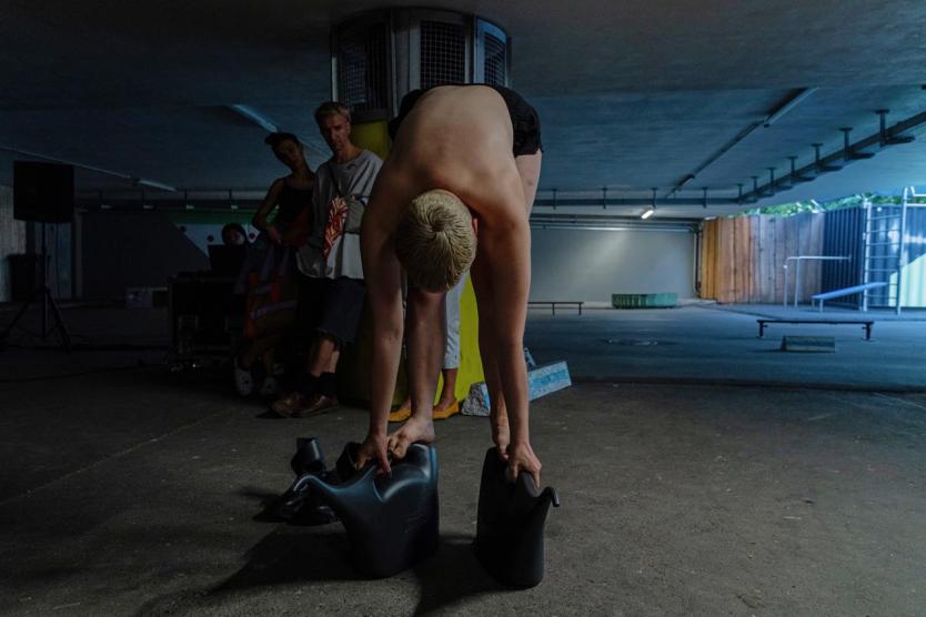 Vor einem Pfeiler in einem Parkhaus beugt sich ein nackter Oberkörper nach vorn, beide Hände ruhen auf zwei schwarzen Gießkannen, im Hintergrund lehnen drei Personen an der Wand und beobachten die Szene.