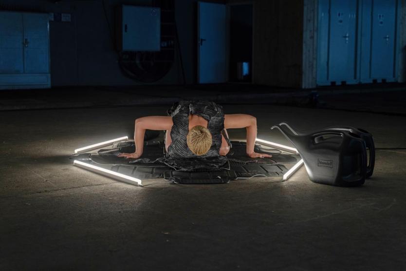 Körper in Bodennähe auf einem Sechseck aus Leuchtstäben und Gummimatten, daneben zwei schwarze Gießkannen auf dem Betonboden eines dunklen Raums.