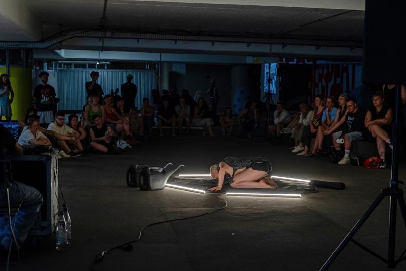 Eine Person kniet auf einer leuchtenden Plattform inmitten eines Parkhauses, umgeben von Kanistern und einem sitzenden Publikum im Halbkreis.