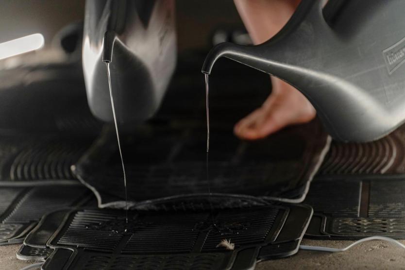 Zwei Gießkannen lassen dünne Wasserstrahlen auf übereinanderliegende schwarze Gummimatten tropfen, im Hintergrund sind nackte Füße sichtbar.