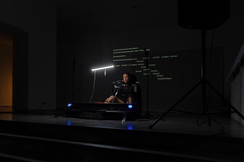 Eine Performer*in sitzt in einem Lenksimulator unter grellem Licht, während Textfragmente mit klimatischen Warnungen an die Wand projiziert werden – eine dichte Szene zwischen Kontrolle, Krise und Konzentration.