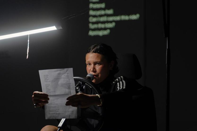 Eine Person im Trainingsanzug sitzt hinter einem Lenkrad, spricht in ein Mikrofon und hält ein beschriebenes Blatt Papier, darüber leuchtet eine Lampe, im Hintergrund erscheinen projizierte Textzeilen.