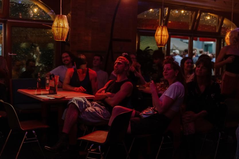 Das Publikum sitzt in einem stimmungsvoll beleuchteten Raum an kleinen Tischen und hört aufmerksam zu. Einige lachen oder lächeln – die Atmosphäre ist locker bei der Stand-up-Performance.