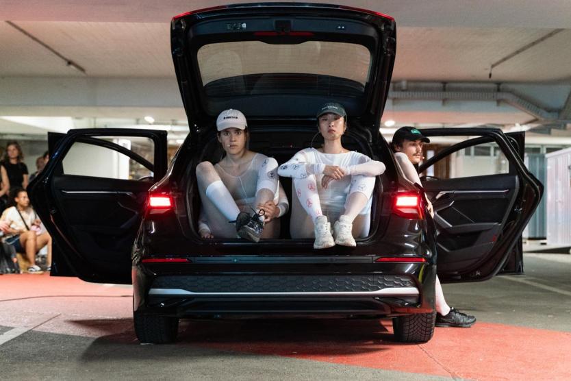 Drei Performer*innen sitzen in einem geöffneten Auto im Parkhaus – ruhige Körperhaltung trifft auf kühle Präsenz inmitten urbaner Kulisse.