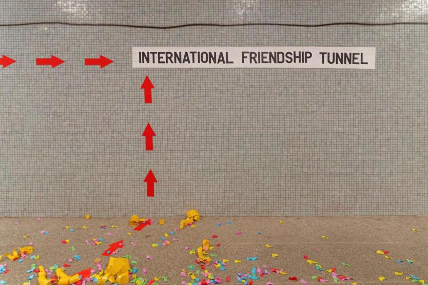 Mosaikfliesenwand mit roter Pfeilmarkierung und dem Schriftzug „INTERNATIONAL FRIENDSHIP TUNNEL“. Auf dem Boden verstreutes buntes Konfetti und Luftschlangen.