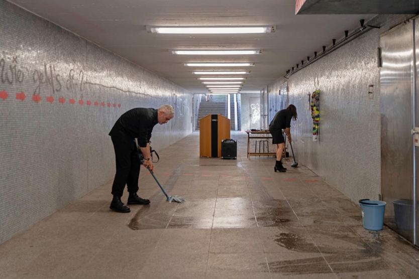 Zwei Personen wischen den Boden eines gefliesten Tunnels. In der Mitte steht ein Rednerpult, an den Wänden sind Kunstwerke und rote Pfeile angebracht. Der Raum wirkt wie eine vorbereitete oder nachbereitete Veranstaltungsfläche.