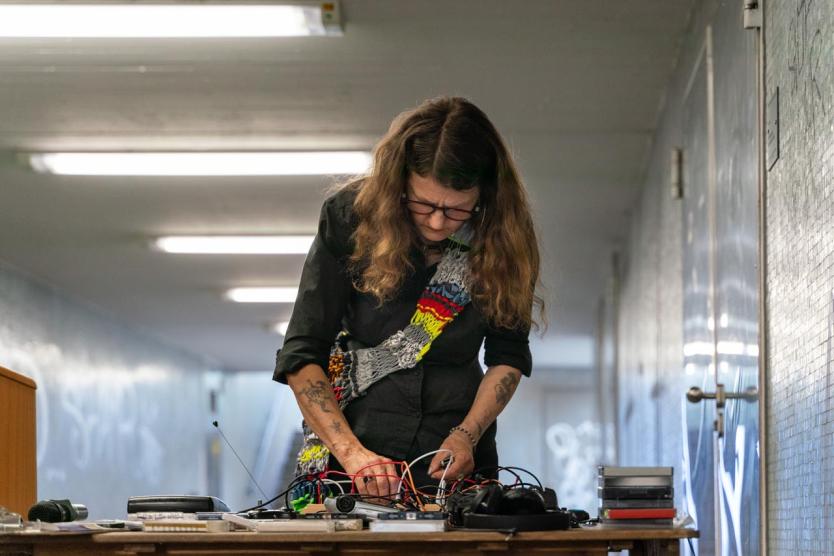 Person mit langem Haar steht an einem Tisch in einem Tunnel und arbeitet konzentriert mit Kabeln und Technik. Der Tisch ist voll mit Geräten, Kassetten und Mikrofonen. Die Umgebung ist gefliest und grafittibeschrieben.