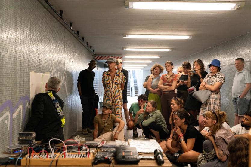 In einem Tunnel versammelt sich eine Gruppe um eine Wandzeichnung, die von einer Person erklärt wird. Umgeben von Technik und Kassetten lauschen die Anwesenden aufmerksam der Präsentation.