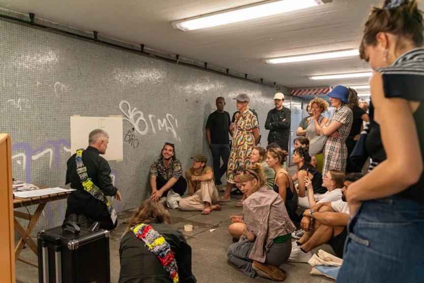 In einem Tunnel versammelt sich eine Gruppe um eine Wandzeichnung, die von einer Person erklärt wird. Umgeben von Technik und Kassetten lauschen die Anwesenden aufmerksam der Präsentation.