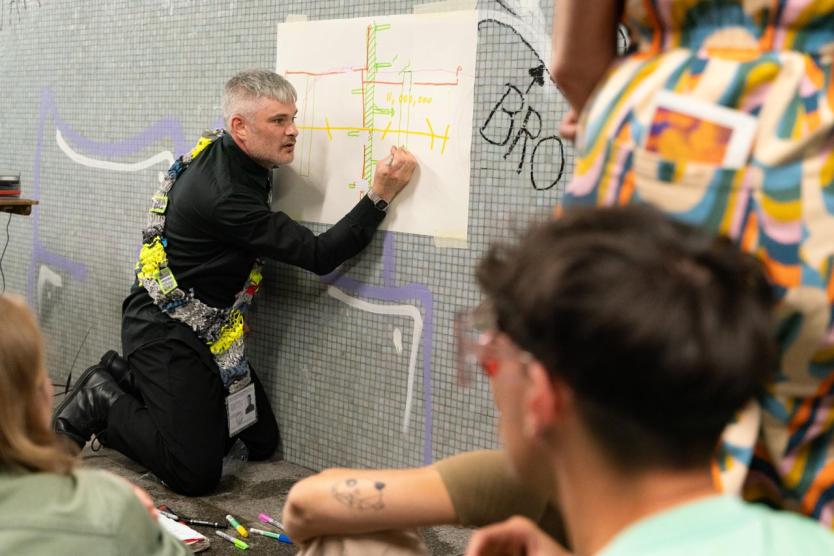 Eine Person kniet an der Tunnelwand und zeichnet mit bunten Markern ein schematisches Diagramm auf ein Blatt Papier. Sie trägt ein gehäkeltes Band mit Namensschild – vermutlich Teil einer Performance oder Präsentation. Umstehende Personen verfolgen aufmerksam das Geschehen.