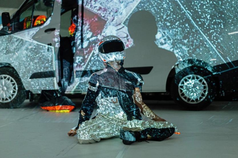 Lichtprojektionen treffen auf eine sitzende Figur in Motorradkleidung vor einem geöffneten Transporter – eine Verschmelzung von Körper, Fahrzeug und digitaler Bildwelt.