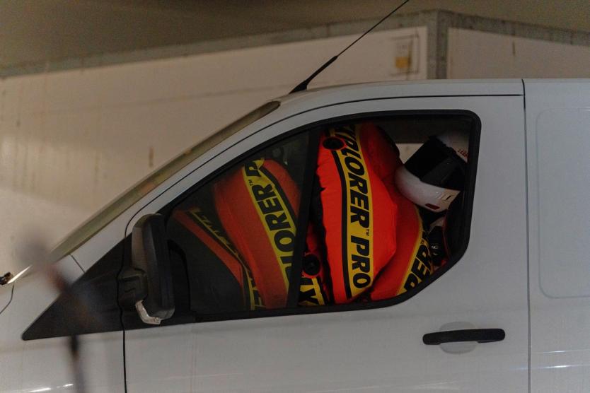 Ein Transporter mit überfüllter Fahrkabine, in der leuchtend orange Schwimmreifen und ein Helm eine absurde Verdichtung von Raum, Sicherheit und Mobilität inszenieren.