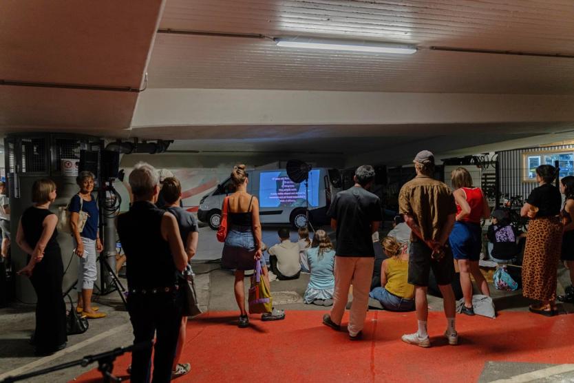 Publikum im Parkhaus versammelt sich vor einem Transporter mit Karaoke-Projektion – eine performative Intervention im urbanen Raum.