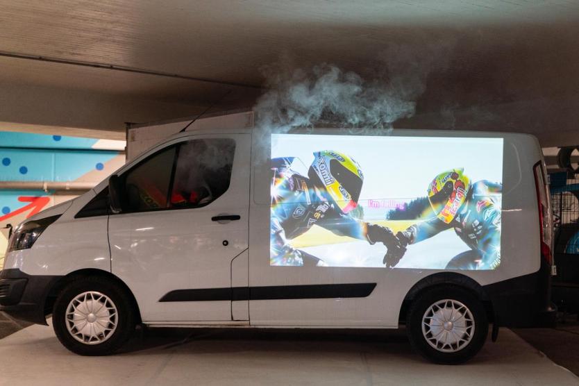 Ein weißer Transporter in einem Parkhaus mit dampfgefüllter Fahrerkabine, auf dessen Seitenwand eine Video-Projektion zweier sich berührender Motorradfahrerhelme mit dem Karaoke-Text „I'm falling“ zu sehen ist.