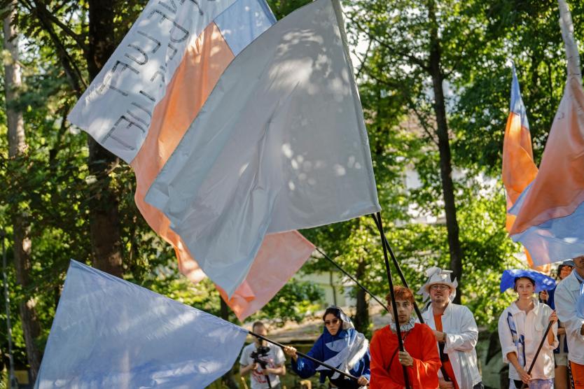 Mehrere Menschen tragen große Fahnen in Weiß, Blau und Orange durch einen sonnigen, baumbestandenen Park. In farbenfroher, teils künstlerischer Kleidung nehmen sie offenbar an einer Performance oder einem Umzug teil.