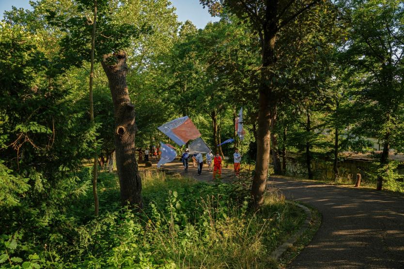 Eine Gruppe bunt gekleideter Menschen zieht mit großen, farbigen Fahnen einen schattigen Waldweg entlang. Die Szene wirkt wie ein künstlerischer Umzug oder eine Performance im öffentlichen Raum.