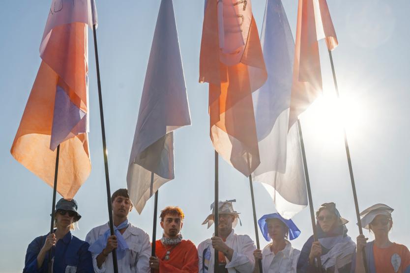 In der tief stehenden Sonne stehen mehrere Menschen in farbenfroher Kleidung mit großen Fahnen in Orange, Weiß und Blau aufgereiht nebeneinander. Die Szene wirkt wie ein feierlicher Moment innerhalb einer künstlerischen Aktion im Freien.