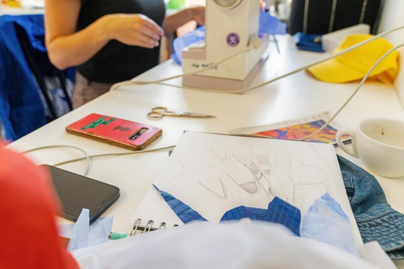 Auf einem Arbeitstisch im Atelier liegen ein Skizzenbuch mit Zeichnungen, Stoffreste, zwei Handys, eine Schere und eine benutzte Kaffeetasse. Im Hintergrund wird an einer Nähmaschine gearbeitet.