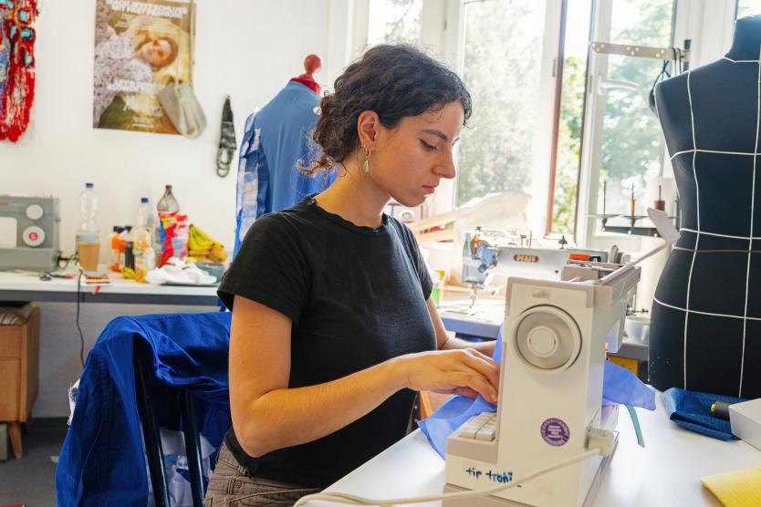 In einem Atelier sitzt eine Person an der Nähmaschine und arbeitet konzentriert mit blauem Stoff. Umgeben ist sie von Schneiderpuppe, Werkzeugen und bunten Materialien.