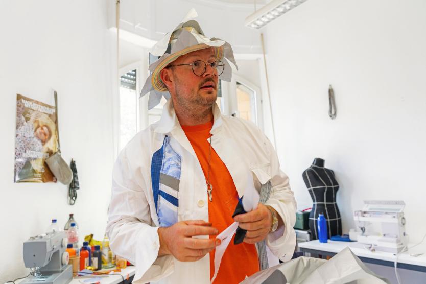 In einem kreativen Atelier hält eine Person Stoffstücke in der Hand. Mit auffälligem Hut und bunter Kleidung steht sie zwischen Nähmaschinen und Materialien.