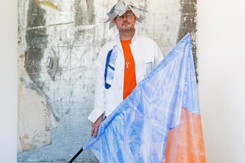 Vor einer abstrakten, silber-grauen Bildfläche steht eine Person mit großer orange-blauer Fahne. Die Kleidung kombiniert Weiß und kräftiges Orange, ergänzt durch einen auffälligen, skulpturalen Kopfschmuck aus Stoff.