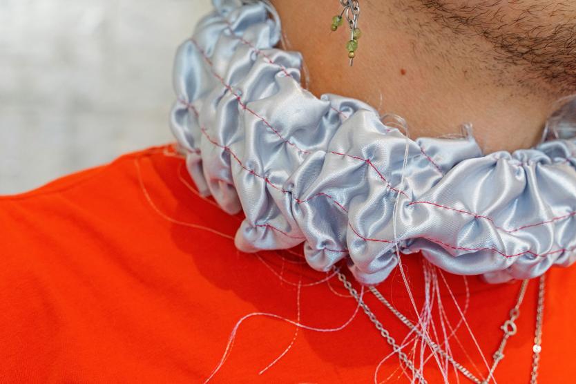 Detailaufnahme eines kunstvoll gestalteten Outfits mit silbern glänzendem, gekräuseltem Kragen, feinen Fäden und mehreren Ketten mit Anhängern. Ergänzt wird das Outfit durch einen filigranen Ohrring mit grünen Perlen.