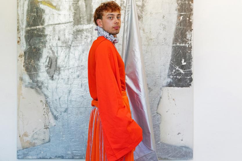 Barfuß vor einer großflächigen, silbrig-schimmernden Wand steht eine Person in einem leuchtend orangefarbenen Gewand mit langen Stofffransen. Eine silberne Fahne lehnt an der Wand und ergänzt die farbliche Inszenierung der Szene.