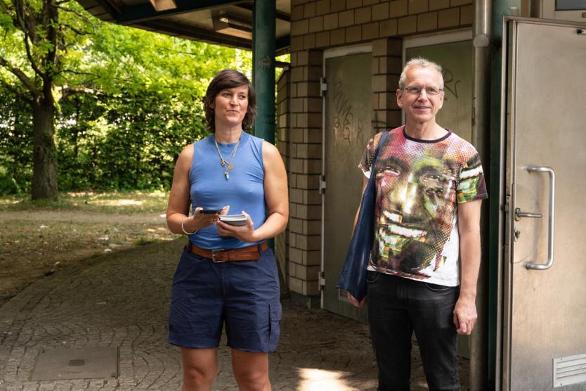 Zwei Personen stehen nebeneinander vor einem öffentlichen Gebäude im Grünen. Sie wirken vorbereitet und zugewand.