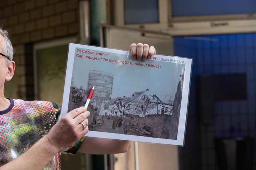 Eine Person hält eine historische Luftaufnahme mit der Aufschrift „25.02.1944 – THE DAY THE CLOUDS VANISHED“. Auf der Karte sind Orte wie „Robert Bosch Hospital“ und „Our Walk“ markiert. Die Person zeigt mit einem silber-roten Stift auf einen Punkt.