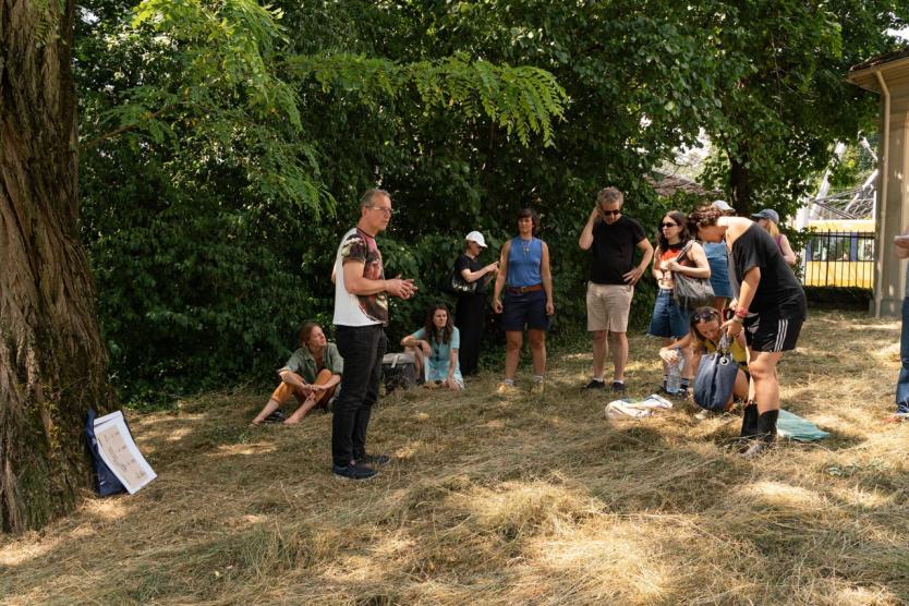 Eine Gruppe versammelt sich auf einer Wiese im Schatten großer Bäume. Eine Person spricht, andere sitzen oder stehen aufmerksam, Materialien liegen auf dem Boden.