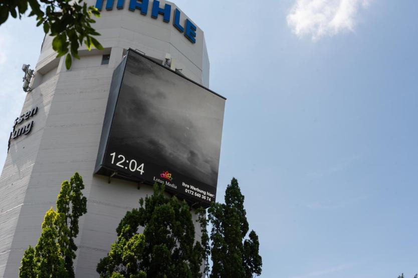 Großer Werbeturm mit digitaler Anzeige zeigt ein düsteres Schwarz-Weiß-Bild von Wolken, darunter steht die Uhrzeit „12:04“. Umgeben von grünen Bäumen und blauem Himmel.