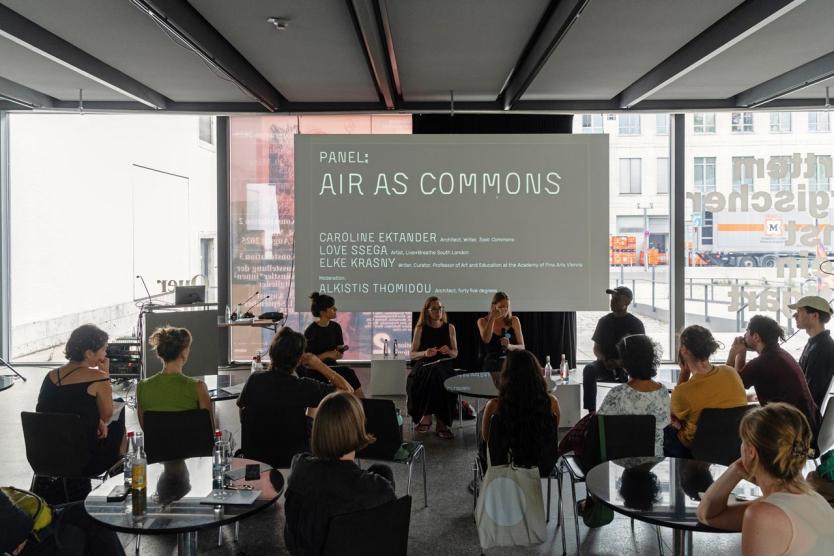 Eine Podiumsdiskussion mit vier sprechenden Personen vor einer großen Projektionsfläche mit dem Titel „Air as Commons“. Das Publikum sitzt verteilt im Raum und hört aufmerksam zu. Helle Fenster im Hintergrund lassen Tageslicht herein.