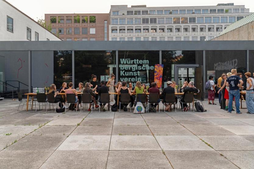 Eine Gruppe von Menschen sitzt bei einem sommerlichen Abendessen an einer langen Tafel im Freien.