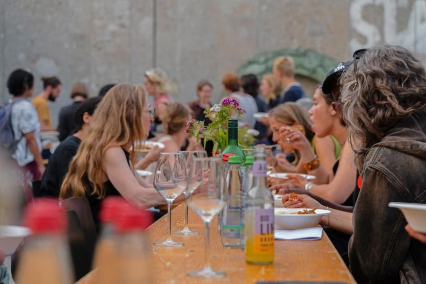 Menschen essen und unterhalten sich an einer langen, sommerlich gedeckten Tafel im Freien. Im Vordergrund stehen Gläser und Flaschen, in der Mitte bunte Blumen. Die Atmosphäre ist lebendig und gesellig.