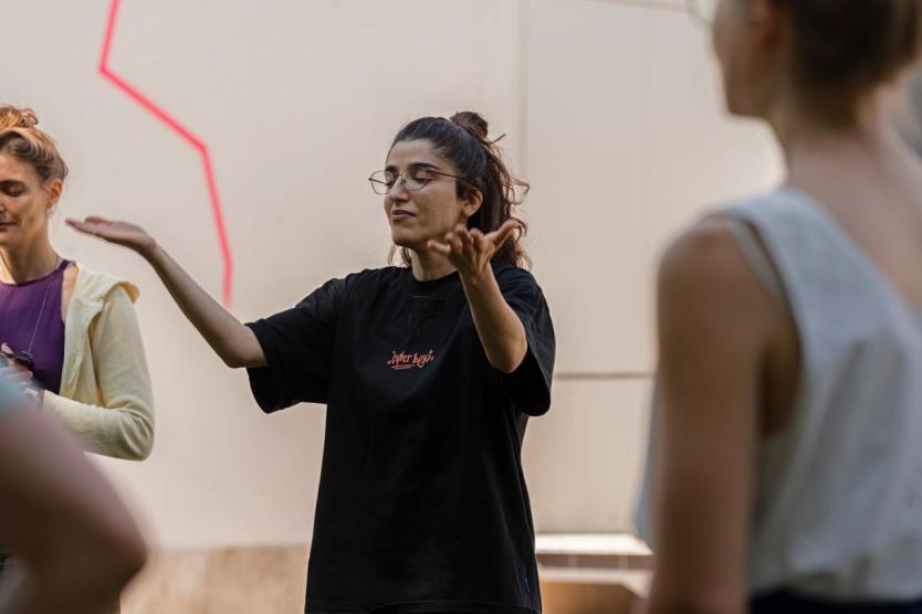Eine Person mit Brille und schwarzem T-Shirt steht mit erhobenen Armen inmitten einer kleinen Gruppe. Sie scheint ruhig zu sprechen oder zu erklären. Im Hintergrund ist eine weiße Wand mit roter Linie sichtbar.
