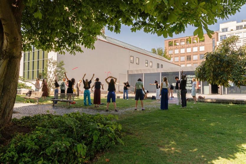 Eine Gruppe an Personen steht im Gras unter Bäumen in einem Kreis und eine Person leitet eine Entspannungsübung an. Die Sonne scheint.