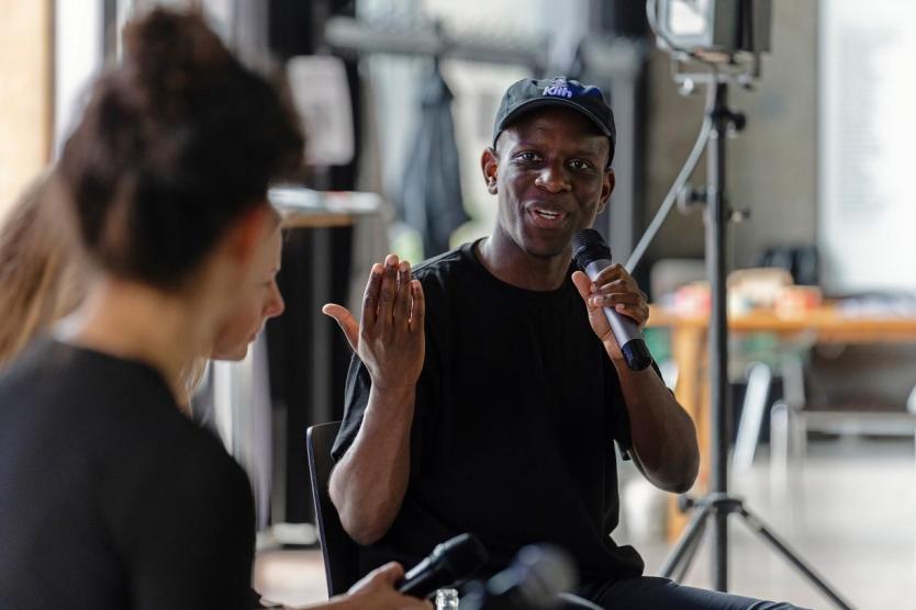 Eine Person in schwarzem T-Shirt und Baseballcap spricht in ein Mikrofon. Die Person sitzt in einem hellen Raum und gestikuliert engagiert mit der freien Hand, während eine andere Person ihm aufmerksam zuhört.