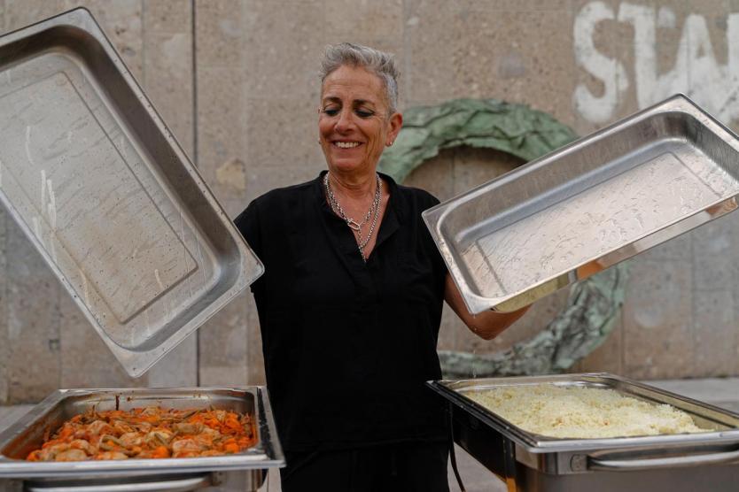 Person mit kurzen grauen Haaren und schwarzer Kleidung steht vor einer Steinwand und hält zwei geöffnete Edelstahlbehälterdeckel in den Händen. In den Warmhaltebehältern befinden sich links ein Gericht mit Gemüse und rechts eine große Menge Reis.