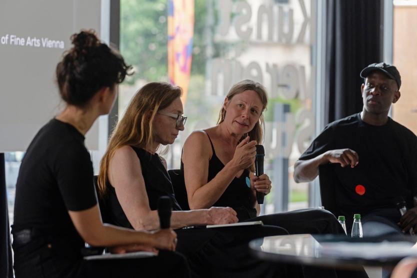 Vier Personen sitzen in einer Gesprächsrunde auf einer Bühne. Eine Frau mit langen blonden Haaren spricht gerade ins Mikrofon, während die anderen aufmerksam zuhören. Die Atmosphäre wirkt konzentriert und ernst.