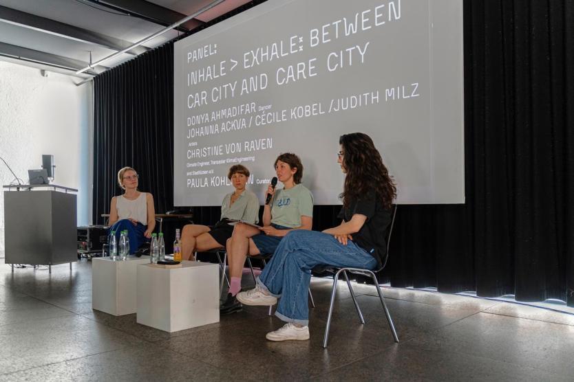 Vier Frauen sitzen auf einer Bühne vor einem großen Projektionstext. Eine spricht ins Mikrofon, die anderen hören aufmerksam zu. Die Szene wirkt ruhig und konzentriert – ein moderiertes Gespräch in einem hellen Raum.