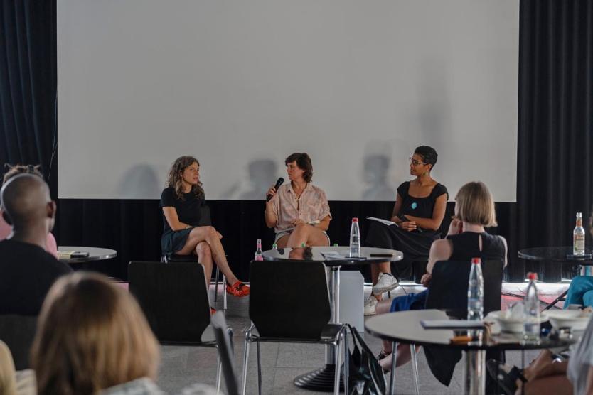 Drei Personen sitzen auf einer kleinen Bühne vor einem weißen Hintergrund und diskutieren mit Mikrofonen. Sie sprechen engagiert, das Publikum hört aufmerksam zu. Die Szene wirkt ruhig und konzentriert.