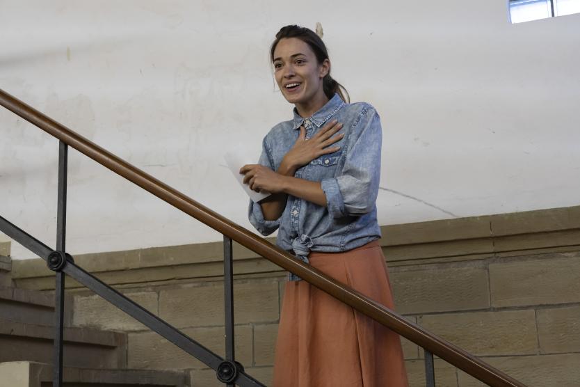 Das Bild zeigt eine Person mit locker gebundenem Dutt auf Treppe, gekleidet in Jeanshemd und orangefarbenem Rock, mit Zettel in einer Hand und anderer Hand auf Brust
