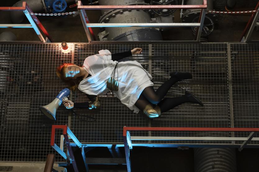 Das Bild zeigt eine Person in weißem Gewand mit schwarzen Strümpfen und goldfarbenen Schützern, liegend auf Gitterboden zwischen Industrieanlagen, mit Megafon in der Hand und buntem Licht angestrahlt.