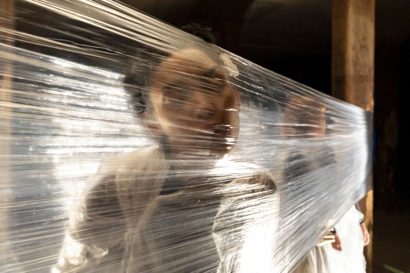 Das Bild zeigt eine Person mit Gesicht dicht an gespannter transparenter Folie, durch die Licht fällt und Konturen verzerrt, während Hintergrund dunkel bleibt.